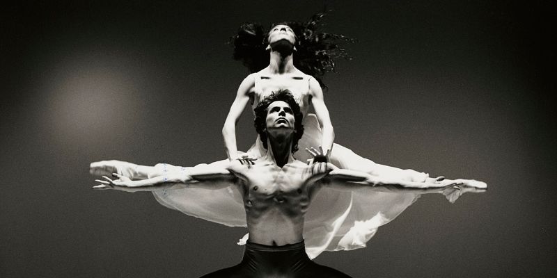 Jayne Regan Pink as Juliet balances with her hands on the shoulders of William Walker as Romeo. Pink holds her legs out and Walker holds his arms out so they form a figure like a cross.