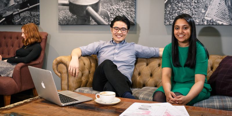 Two postgraduate students sitting on a sofa, working on a laptop and looking at papers together. They are smiling.