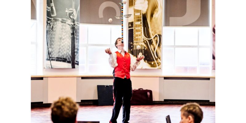 Male circus performer balancing a long multi-coloured stick with three sieves. He is also juggling three white balls