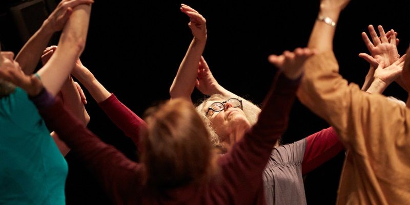A group of people in colourful shirts are facing each other in a circle, reaching their arms upwards as if in the middle of a dance of theatrical performance