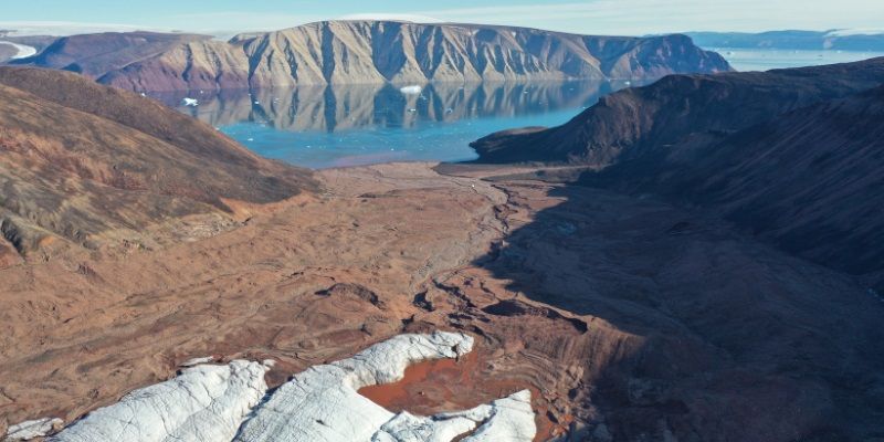 A beautiful view of Greenland showing a small patch of snow in the foreground against a backdrop of greenery, with a lake in the background