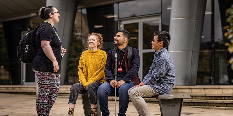 A group of students are chatting with University support staff outside a building. Three are sat on a bench, one person is standing. One of the seated students has a walking cane.