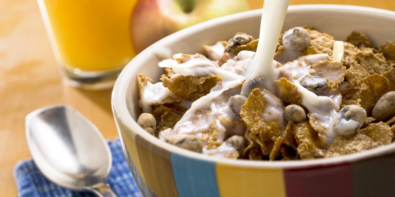 Milk being poured on a bowl of cereal, with an apple and glass of orange juice alongside.