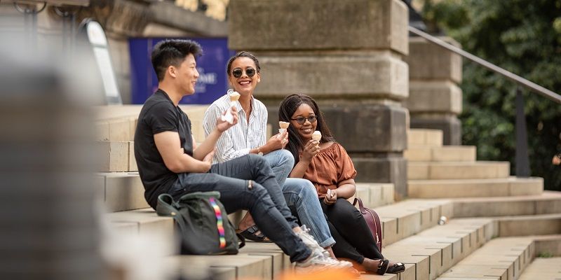 A group of students sat on some steps outside the Leeds City Museum on a sunny day, chatting and eating ice cream