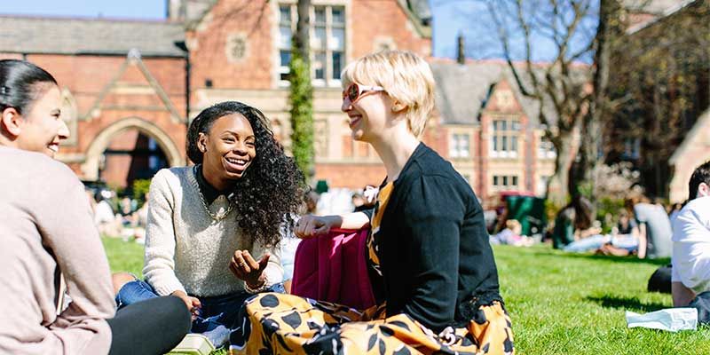 Three students sitting talking on the grass, outside the Great Hall, on campus.