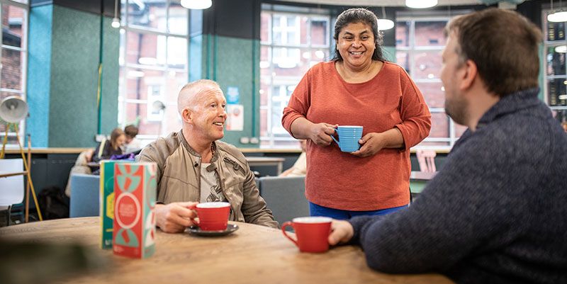 Three people chatting an drinking coffee in a cafe. Two are sitting and one is standing