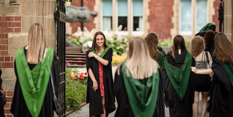 Graduates in their gowns on graduation day. One graduate is facing the camera smiling and wearing a red dress and a gown