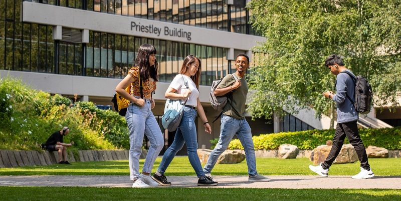 Three students talking and walking across university campus on a sunny day with Priestley Building in the background