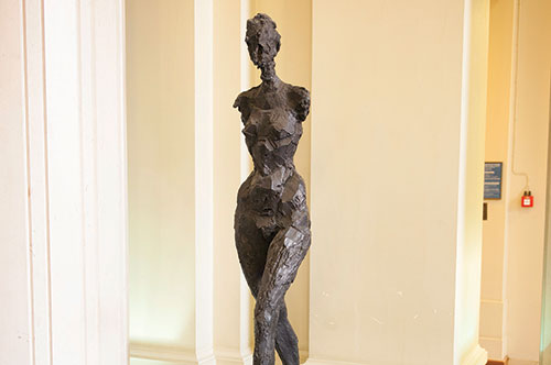 Walking Figure, a sculpture of a female form without arms made of roughly-textured bronze, stands in front of an interior wall.