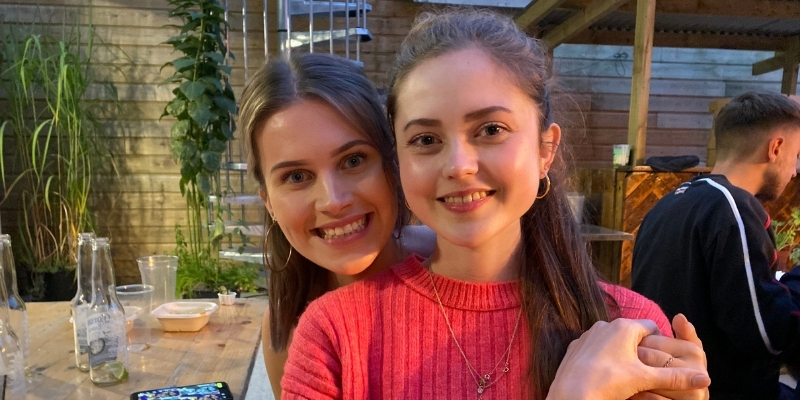 Two young women smile at camera whilst sitting together on a bench and holding hands