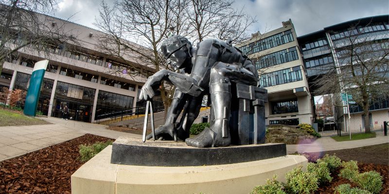 The 'Master of the Universe' sculpture from the side, with the Edward Boyle Library in the background.