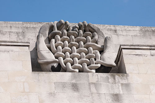 Manmane Fibres, pictured high up on the Clothworker's Building, shows two hands holding woven material. The sculpture is made from Portland stone and blends with the building, as if part of it.
