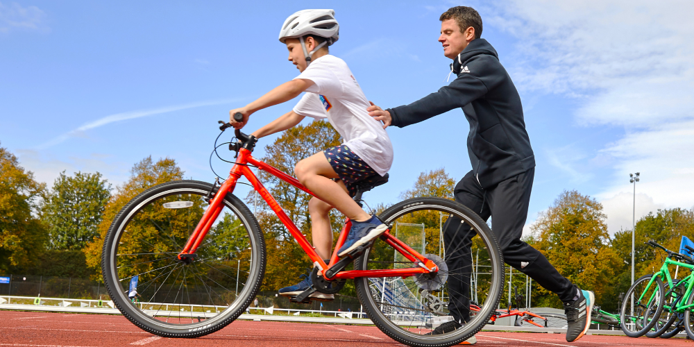 Jonny Brownlee pushes a child along on a bicycle, supporting them as they set off