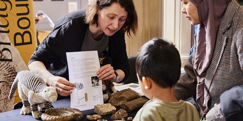 A staff member at Be Curious Live is behind a table with fossils on it. They are talking to a small child and their parent and showing them some information.