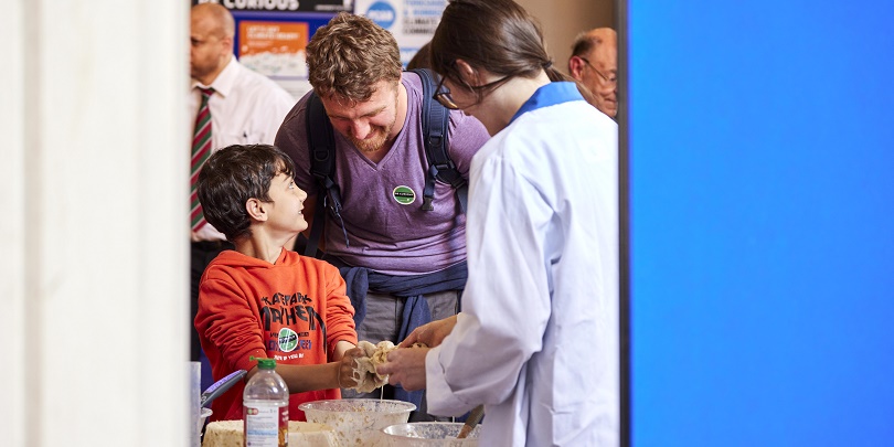 A child and their parent are taking part in an activity to mix up some substances in a bowl with a staff member at be Curious Live. The child now has messy hands and is smiling.