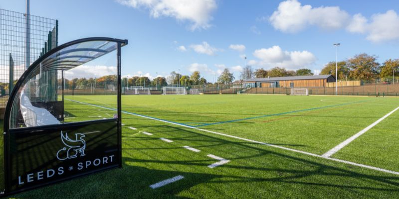 A ground-level photo of a football pitch with a sign that says 'Leeds Sport', and a blue sky.