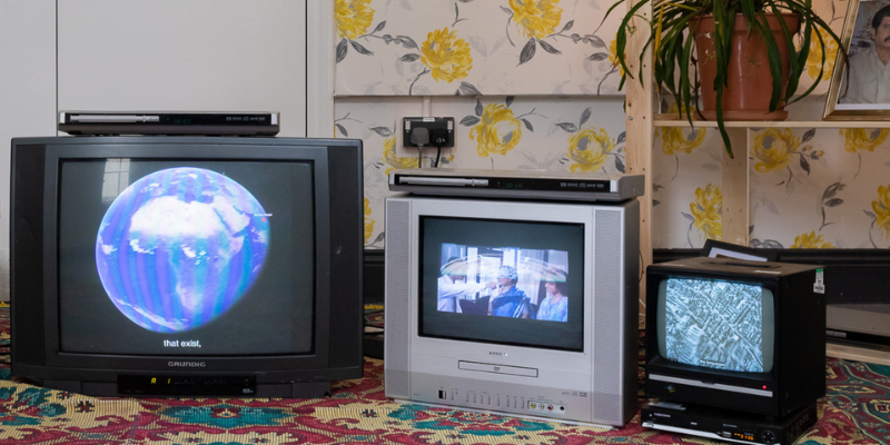 Suman Shams’ art installation. Three retro televisions are shown on a carpeted floor in a household setting. The televisions are showing various videos of the globe and famous people.