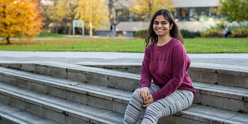 Hetasha Gopalani sitting on some stone steps. There is grass and trees with orange and yellow leaves in the background.