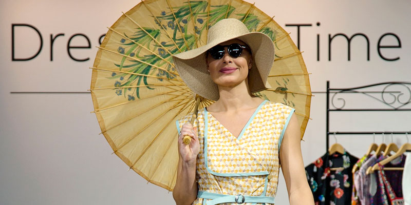 A person wearing a yellow dress, hat and umbrella at a fashion show.