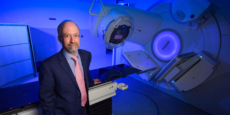 Professor David Sebag-Montefiore standing next to a radiotherapy machine in a blue room treatment room in a hospital in Leeds.