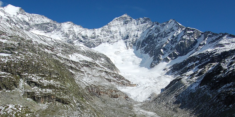 Odenwinkelkees glacier 
 with bare rocky mountainside on either side with bright blue sky in the background. 