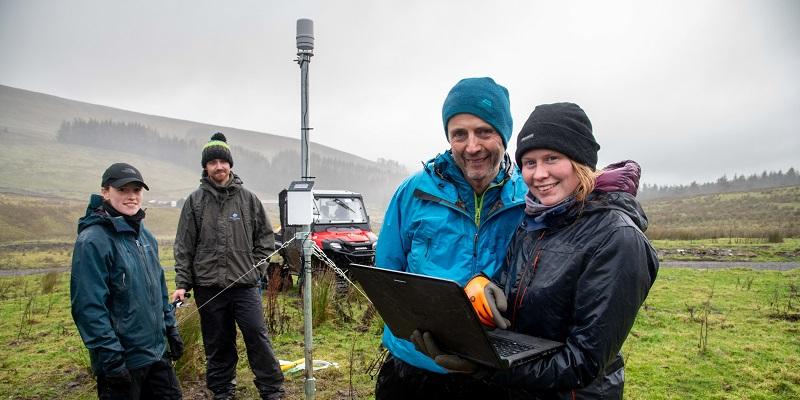 Four scientists are standing near an instrument that captures weather data, which has been installed on open moorland.