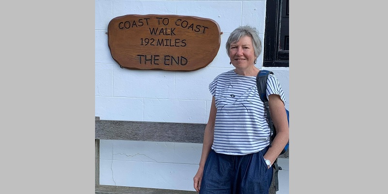 62 year old woman with grey hair in a white and black striped t-shirt stands in front of a white wall with a mounted wooden sign that says Coast to Coast walk, 192 miles, The End.