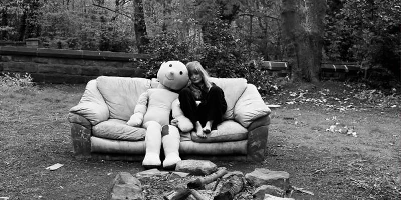 Black and white image of a woman and a large doll on a sofa outside