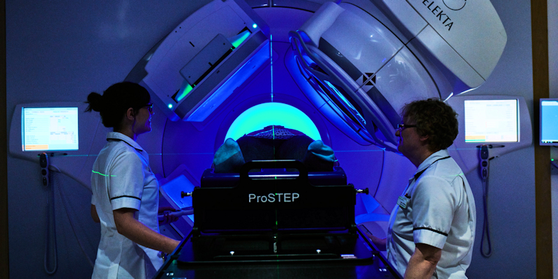 Two nurses stand in a hospital room with a large radiotherapy machine pointing towards a treatment table, under low blue lighting.