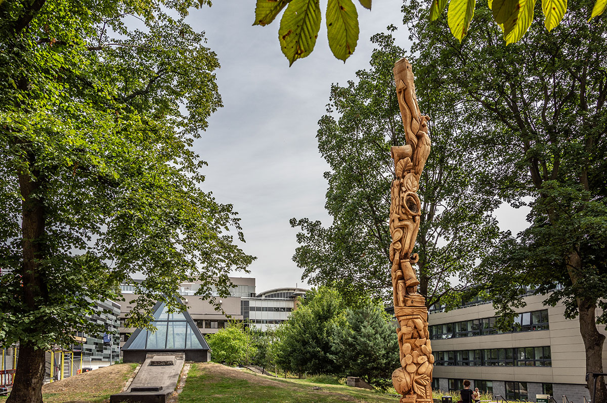 A tall tree carving with intricate designs stands among trees and buildings.