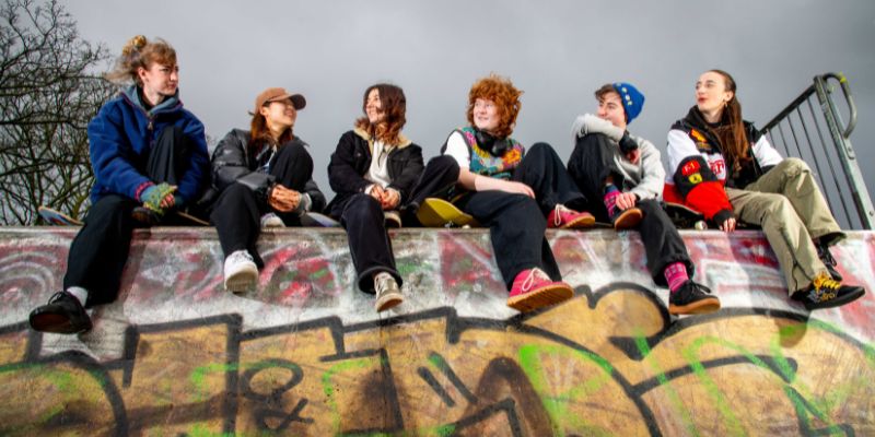 Six young women sit on the top of the skate ramp, talking and smiling, in front of a dark clouds in the sky