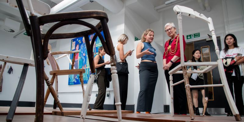 An image of two wooden cafe chairs in a large artworks, as the Lord Mayor of Leeds Al Garthwaite, wearing her gold chains of office, chats to a student in the background.