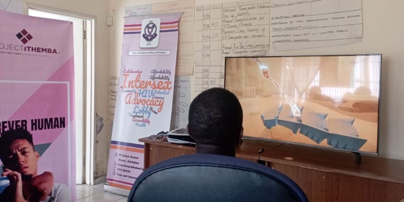 A member of Trans and Intersex Rising in Zimbabwe playing a video game set in a barren desert landscape. They are surrounded by positive posters advertising the group.