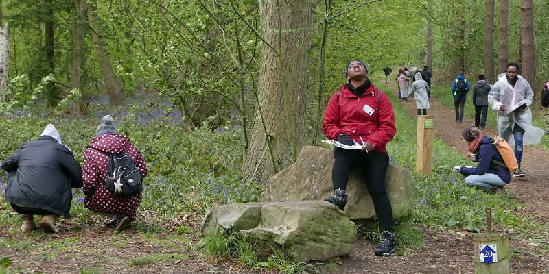 A group of people with clipboard are observing their surroundings in the woods. One women in the foreground is wearing a red coat, she is sitting on a stump looking up at the tree branches