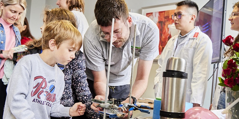 A staff member is demonstrating how to use a small set of engineering equipment to a young child at Be Curious Live. Other people are in the background
