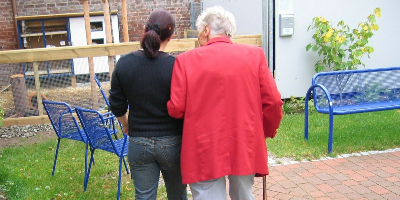 An elderly woman and a care home nurse walking arm in arm, seen from behind