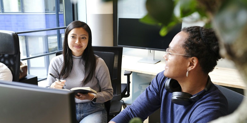Two people sat in a modern bright workspace, looking at computers and chatting.