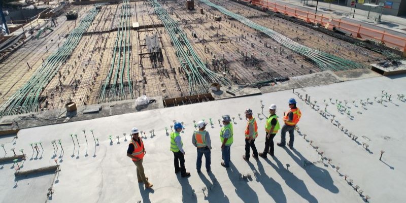 Birds eye view of a row of 7 engineers in hi-vis and hard hats, standing on concrete and looking over a construction site of cables and wires.