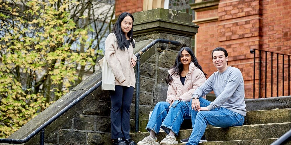 Three students on steps in front of a red brick building. Two are sat on steps, one is standing.
