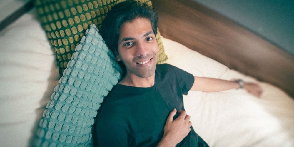 A student lies on their bed, propped up by cushions and pillows. They look up at the camera smiling.