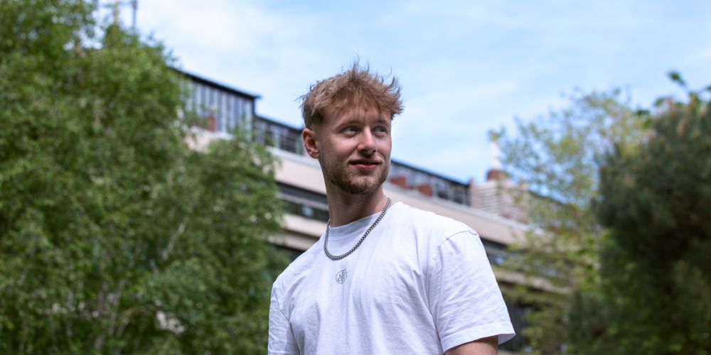 A student on campus on a sunny day with a building and trees in the background.