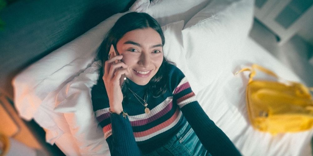Overhead shot of a prospective student sitting on their bed, with their phone to their ear. They are wearing a striped jumper and are looking just past the camera, smiling. A yellow rucksack lies upside-down on the bed next to them.