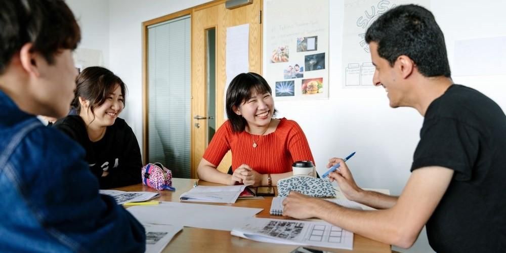 Four students sat around a table smiling. There are papers and pencil cases on the table.