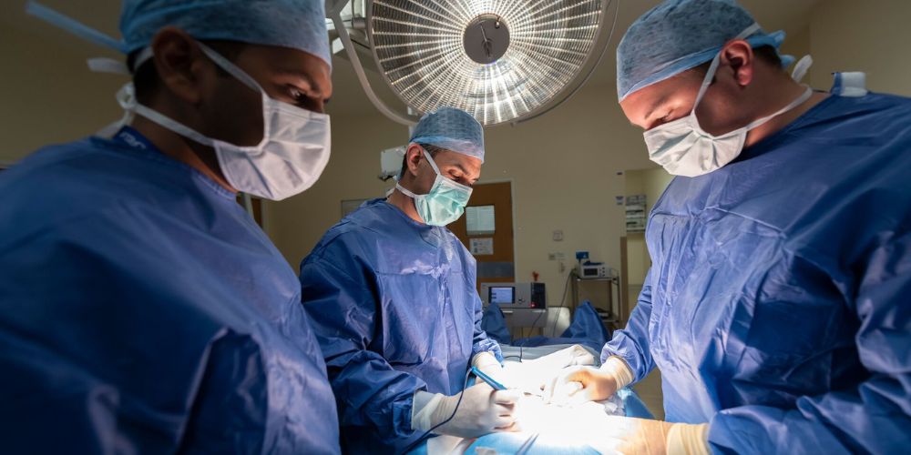A group of surgeons perform a procedure in a sterile operating room. The surgeons are dressed in standard surgical attire, including gowns, masks, gloves, and caps and there is lighting focused on the operating table.