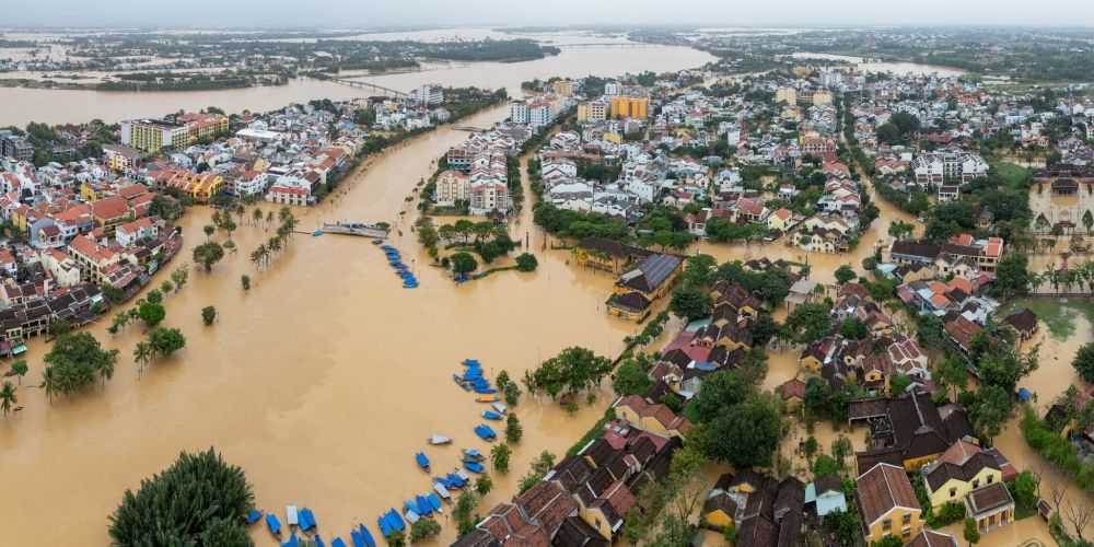 Aerial view of a densely built riverside town with brown floodwaters submerging streets, surrounding houses, and trees, with boats navigating through the inundated area.