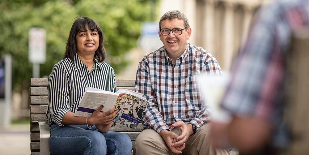 Two mature students sit on a bench outside under a tree, one is holding a textbook