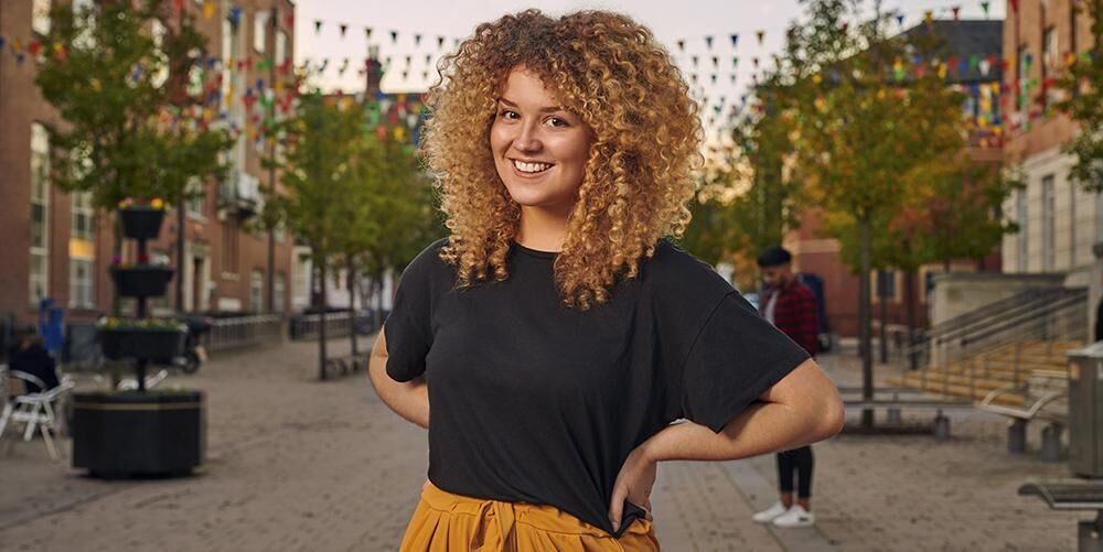 A student wearing a black t-shirt and orange trousers is standing outside Leeds University Union. There are trees and colourful flags in the background.