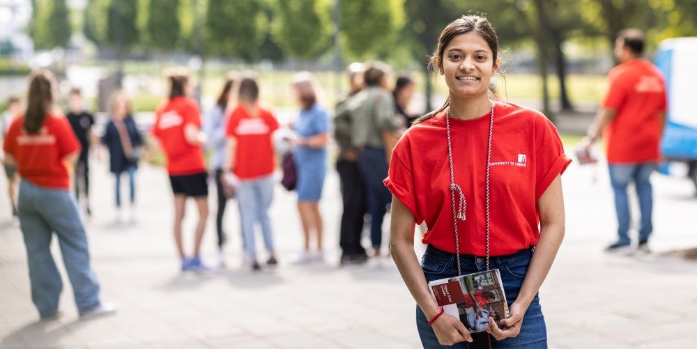 A student ambassador smiling at the camera, with other student ambassadors and visitors in the background, at an open day.