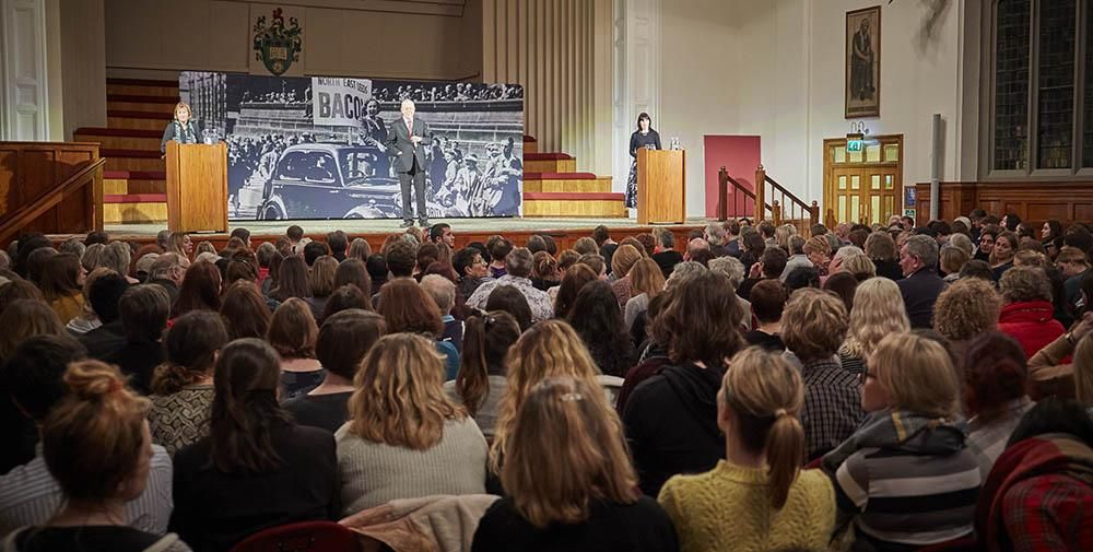 Rows of people sat in front of a speaker on a stage in the Great Hall.