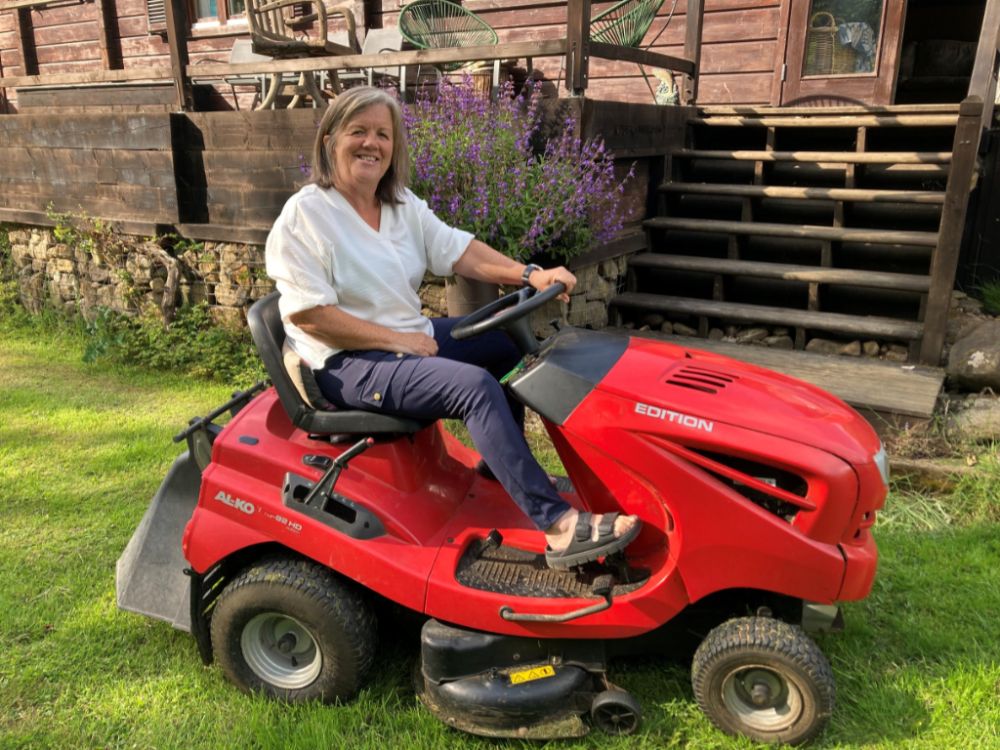 Catherine Whitfield sitting on a red lawnmower, smiling, in her garden